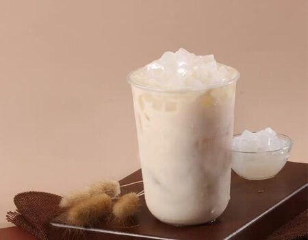 There is a wooden board with a cup of white milk tea placed on it. The top of the milk tea has white coconut jelly, and there is a small bowl of coconut jelly next to it
